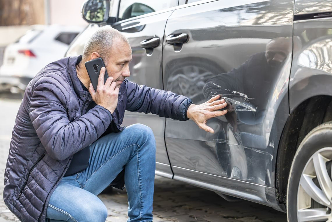 Ein Mann begutachtet Kratzer an einem Auto während er mit seinem Anwalt Verkehrsrecht telefoniert. 